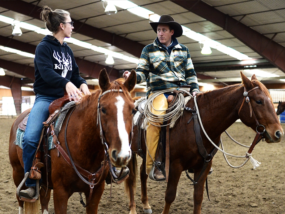 Two riders on horseback discuss technique inside the arena.