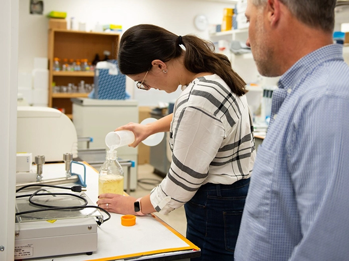 Student pours solution into a flask in a campus lab as an instructor observes.